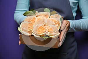 Female florist holding gift box with beautiful roses on color background, closeup