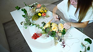 Female florist makes out large floral bouquet of roses with tools, sitting in office in daytime.n