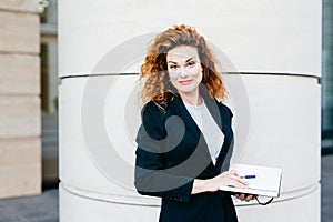 Female freelancer with bushy hairstyle, wearing white blouse, black jacket and skirt, holding her diary book, writing notes in it.