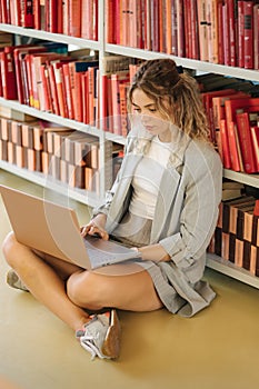 Female freshman sitting on floor in library and study using laptop. Concept of university library