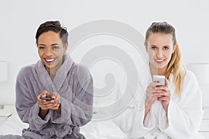 Female friends in bathrobes text messaging on bed