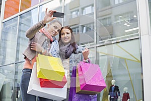 Female friends with shopping bags taking self portrait through mobile phone against store