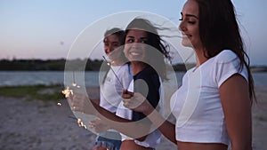 Female friends walking, dancing, having fun at night party at seaside with sparklers in hands. Young teenage women