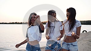 Female friends walking, dancing, having fun at night party at seaside with sparklers in hands. Young teenage women