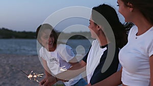 Female friends walking, dancing, having fun at night party at seaside with sparklers in hands. Young teenage women