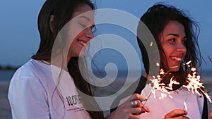 Female friends walking, dancing, having fun at night party at seaside with sparklers in hands. Young teenage women