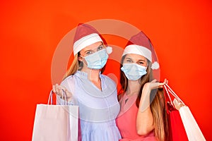 Female friends wearing surgical mask and Christmas hat holding shopping bags on a vivid red orange background