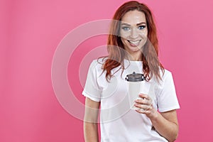 Female with ginger hair using disposable cup of take away coffee, enjoying beverage while holding cup in white casual style shirt