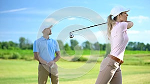 Female golfer hitting ball at course, male coach watching successful shot, sport