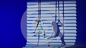 Female gymnast isolated on blue neon studio background. Girl aerial dancer holding and spinning on acrobatic trapeze