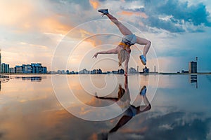 Female gymnast standing on one hand and keeping balance during dramatic sunset with reflection in the water of amazing