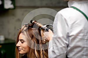 The female hairdresser is curling hair for a brown-haired young caucasian woman in a beauty salon.