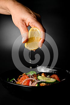 Female hand adds lemon juice to a black bowl with vegetable salad with bread croutons