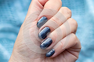 Female hand with beautiful manicure - dark gray blue glittered nails on blue knitted sweater fabric background