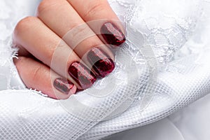 Female hand with beautiful manicure - dark red glittered nails in white satin fabric with lace. Selective focus with