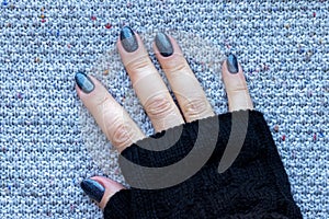 Female hand in black knitted gloves mitts with beautiful manicure - dark blue glittered nails on knitted background