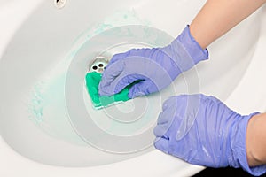 Female hand in a blue glove wipes the sink with a green foam sponge in the bathroom