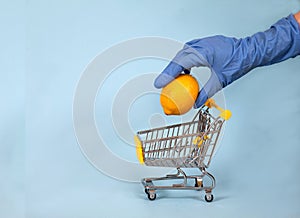 Female hand in a blue rubber glove lowers a lemon into a supermarket trolley on a blue background. Protection. Hygiene. Health.