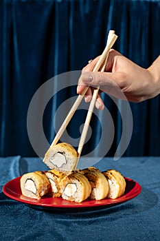 Female hand with chopsticks holding sushi roll on blue velvet background. Roll with salmon on red plate. Modern, stylish
