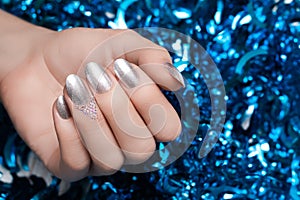 Female hand with Christmas nail design. Silver nail polish manicure. Female hand on blue New Year tinsel background