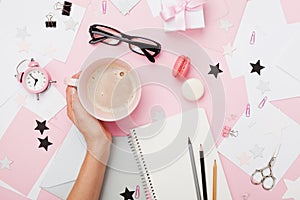 Female hand with coffee cup, macaron, office supply, gift and notebook on pastel desk top view. Fashion pink woman workplace.