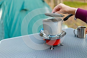 Female hand is cooking in an aluminum pot meals on a portable gas burner on background in the camping in the forest. Women hand ho