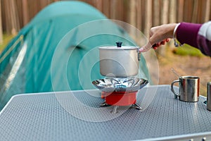 Female hand is cooking in an aluminum pot meals on a portable gas burner on background in the camping in the forest. Women hand ho