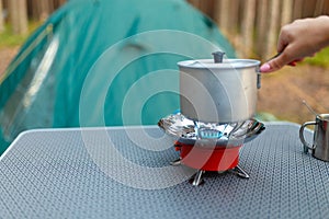 Female hand is cooking in an aluminum pot meals on a portable gas burner on background in the camping in the forest. Women hand ho