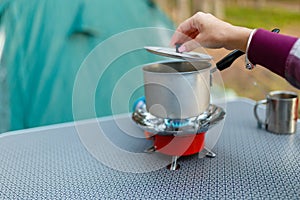 Female hand is cooking in an aluminum pot meals on a portable gas burner on background in the camping in the forest. Women hand ho