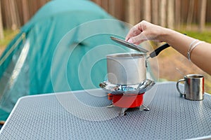 Female hand is cooking in an aluminum pot meals on a portable gas burner on background in the camping in the forest. Women hand ho