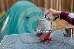 Female hand is cooking in an aluminum pot meals on a portable gas burner on background in the camping in the forest. Women hand ho