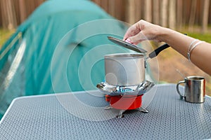 Female hand is cooking in an aluminum pot meals on a portable gas burner on background in the camping in the forest. Women hand ho