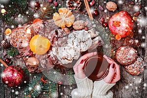 Female hand a cup of tea. Christmas Cookies Chocolate, tea, pomegranate, Tangerines, Nuts, cocoa beans on wooden snowy background.