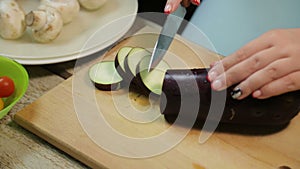 Female hand is cutting a dark eggplant with a knife on a wooden board