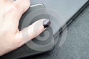 Female hand with dark nails holds a mobile phone with a knitted nylon back, which lies on a closed textured gray laptop cover
