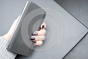 Female hand with dark nails holds a mobile phone with a knitted nylon back, which lies on a closed textured gray laptop cover