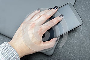 Female hand with dark nails holds a mobile phone, which lies on a closed gray laptop