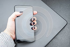 Female hand with dark nails holds a mobile phone, which lies on a closed gray laptop