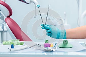 Female hand of dentist in blue gloves holding mouth mirror and periodontal explorer scaler, blurred background of clinic with
