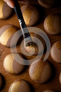 Female hand draws golden stripes on brown macaroons with paint for confectionery products