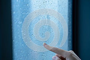 Female hand draws a question mark on a foggy window during the rain. A glass in drops of water