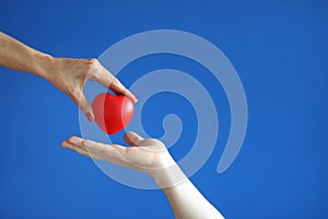 Female hand give red heart to male hand on blue background close-up.