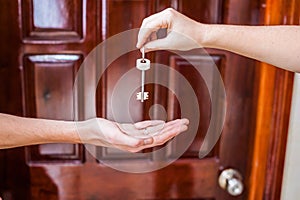 Female hand giving a key to the house man`s on background of wooden door. Owning real estate concept