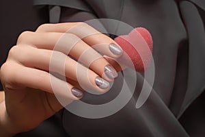 Female hand with glitter beige nail design. Beige nail manicure. Woman hand hold red heart on gray fabric background