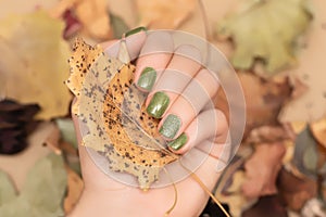 Female hand with green autumn nail design. Woman hand hold dry leaf. Autumn nail polish manicure with golden glitter