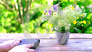 Female hand hold book in black cover, bouquet of wild flowers on wooden table in garden, blurred natural landscape in background