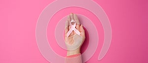 Female hand hold a pink silk ribbon in the form of a loop on a pink background. Symbol of the fight against breast cancer