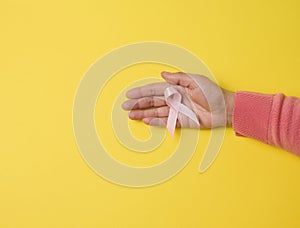 Female hand hold a pink silk ribbon in the form of a loop on a yellow background. Symbol of the fight against breast cancer