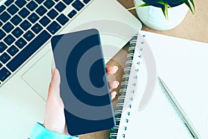 Female hand holding black mobile smart phone with blank screen on laptop computer and notepad background.