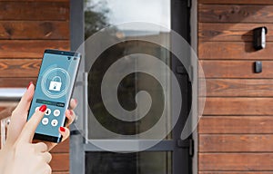 Female hand holding black mobile smart phone with smart home application on the screen. Blurred house on the background